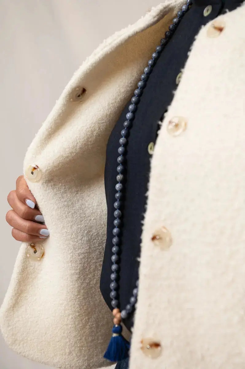 Gilet sans manches Ouessant en drap de laine Casentino crème, vu de profil, détail des boutons effet corne et du côté réversible en jersey de laine crème, porté avec une chemise Narcisse en étamine de laine bleu marine et un collier Mala signé Nadine Tombez.