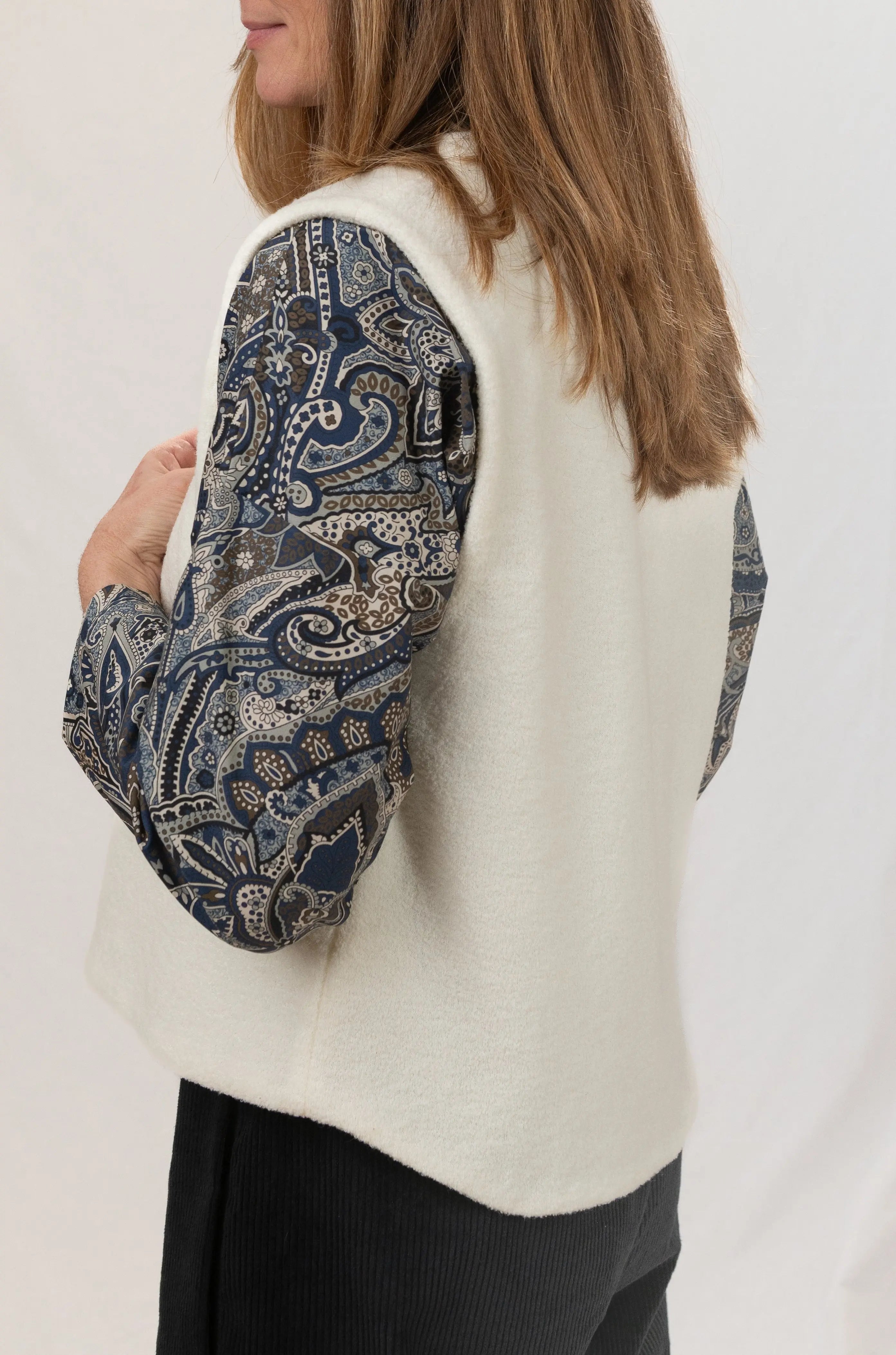 Gilet sans manches Ouessant en jersey de laine couleur crème, vu de profil, réversible, porté avec le pantalon Sylène en velours noir et une chemise Narcisse en popeline de coton à motif paisley cashmere.