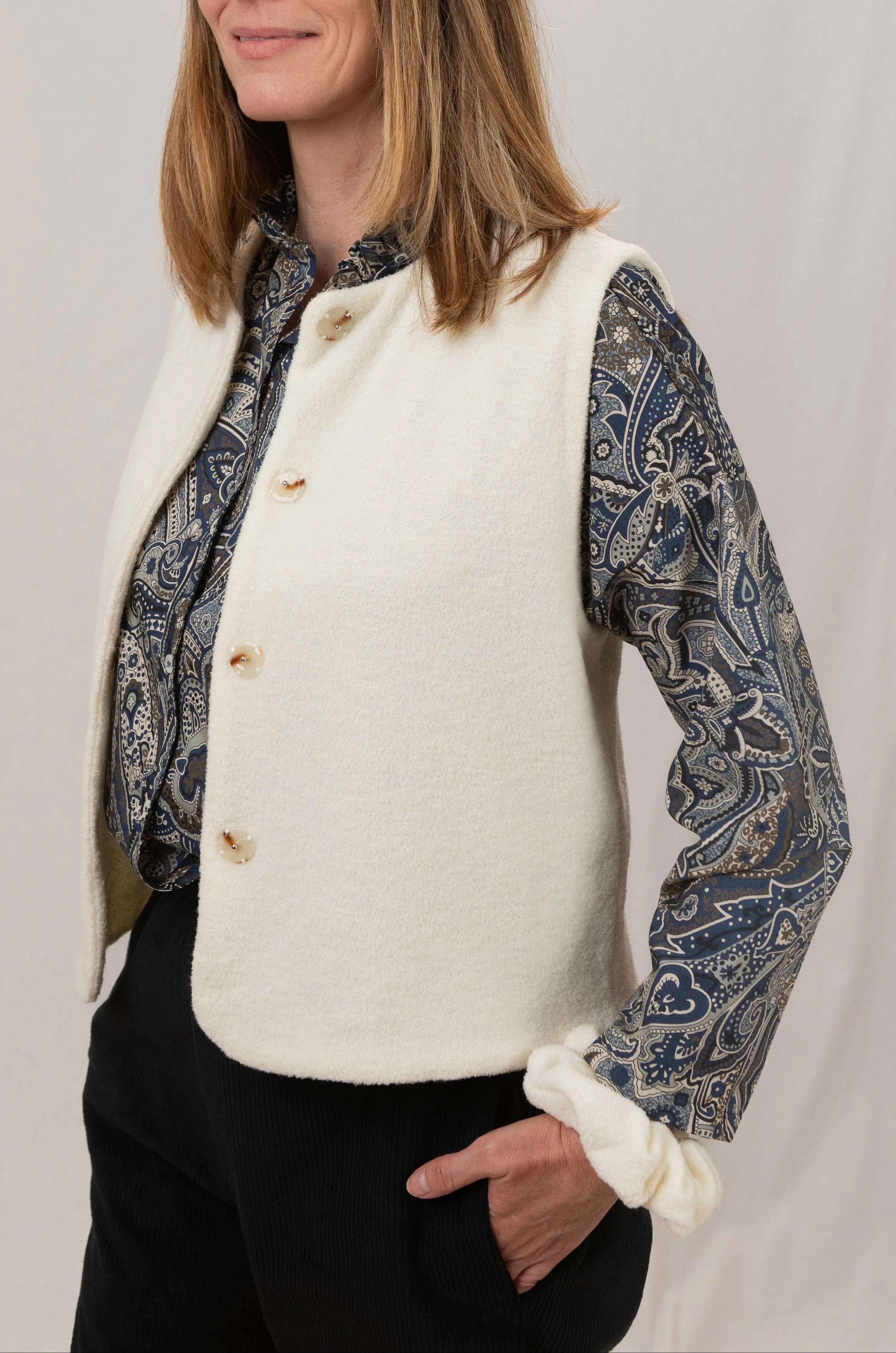 Gilet sans manches Ouessant en jersey de laine couleur crème, vu de profil, réversible, avec boutons effet corne, porté avec le pantalon Sylène en velours noir, une chemise Narcisse en popeline de coton à motif paisley cashmere et un chouchou en jersey de laine crème
