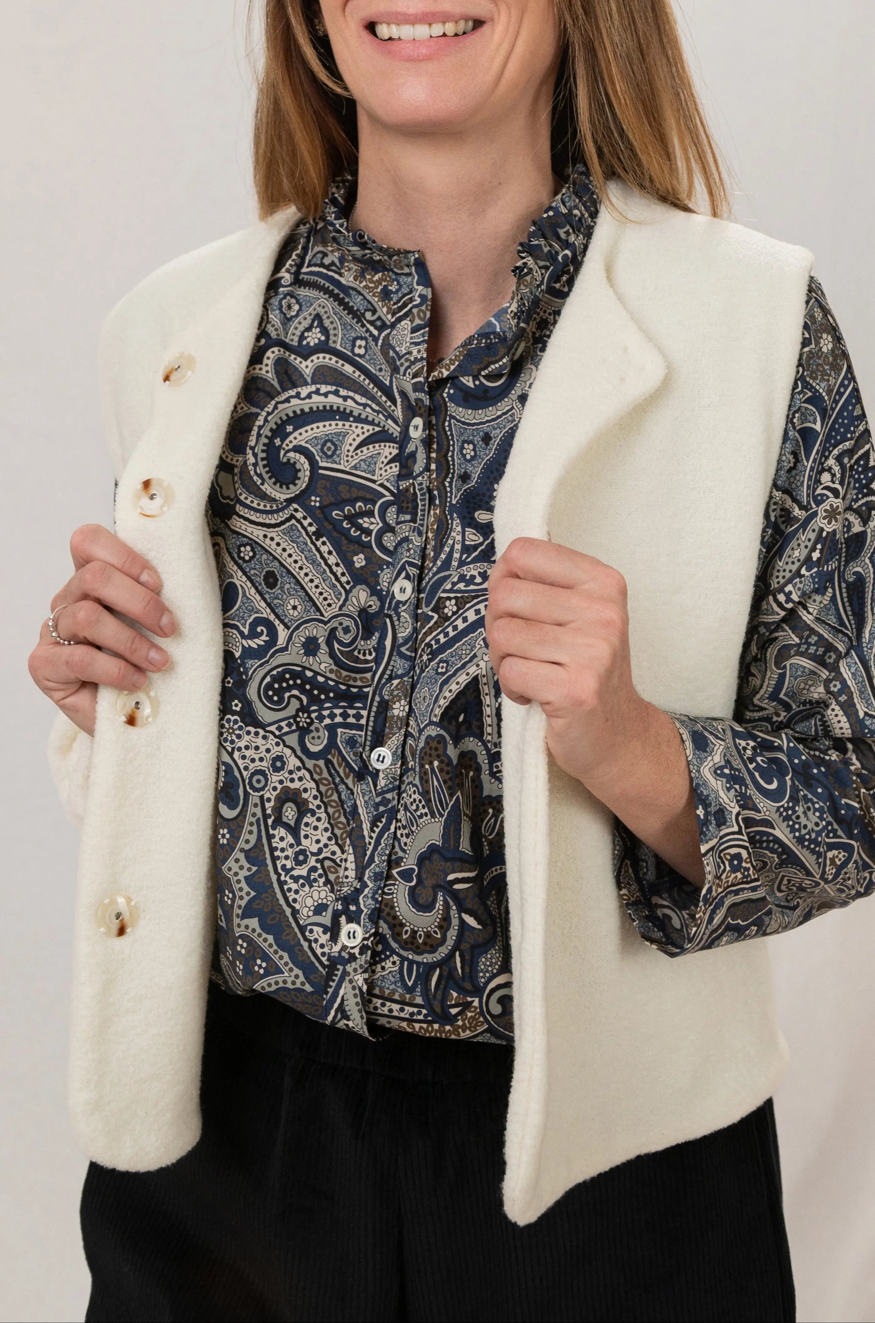 Gilet sans manches Ouessant en jersey de laine couleur crème, réversible, vu de profil, avec boutons effet corne, porté avec le pantalon Sylène en velours noir et une chemise Narcisse en popeline de coton à motif paisley cashmere.
