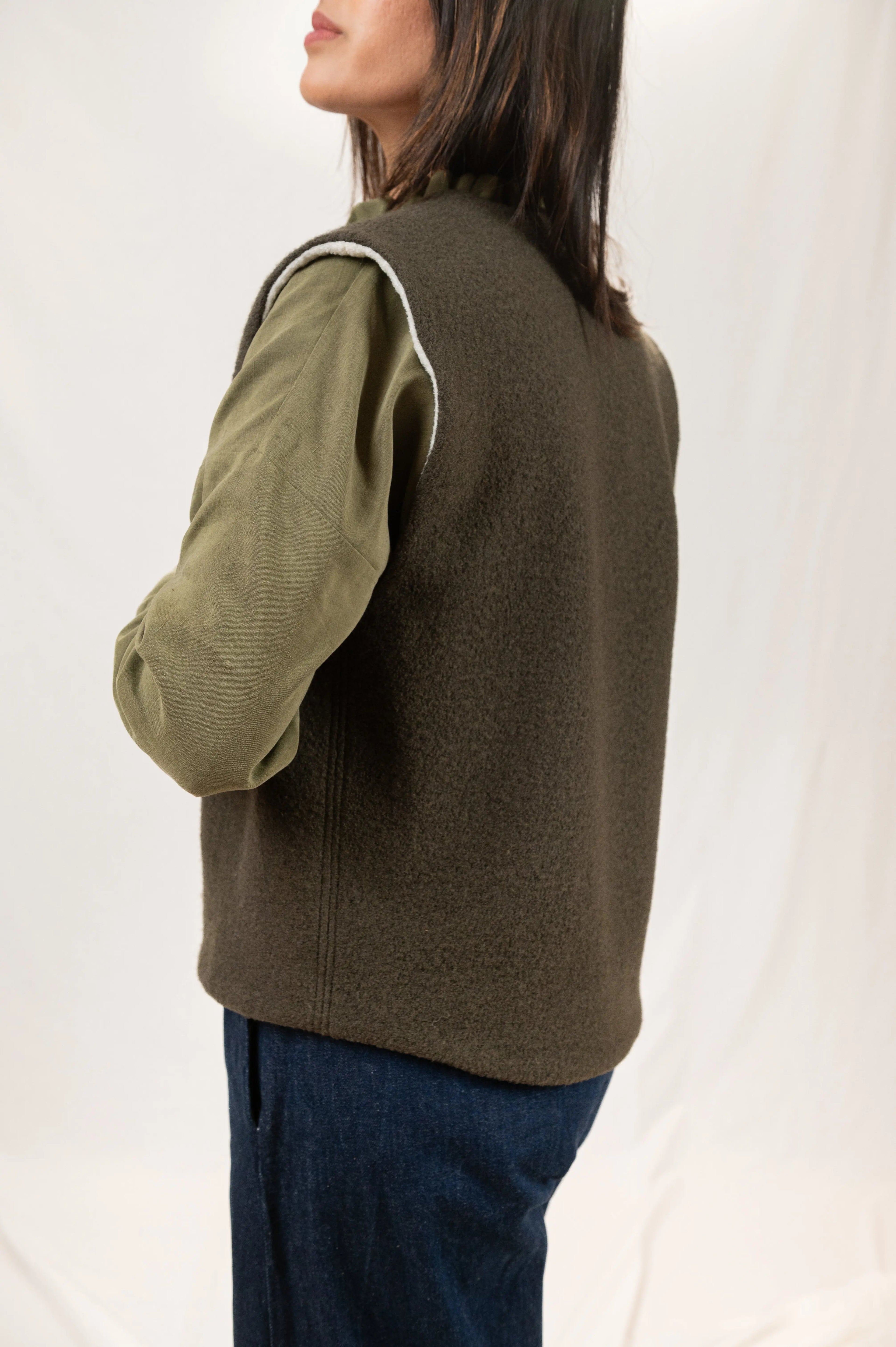 Gilet sans manches Ouessant réversible en laine bouillie vert forêt, vu de profil, détail de la coupe dos recouvrant, porté sur une chemise Narcisse en lin et soie kaki et un pantalon Sylène en denim taille haute, coupe Barrel Leg.