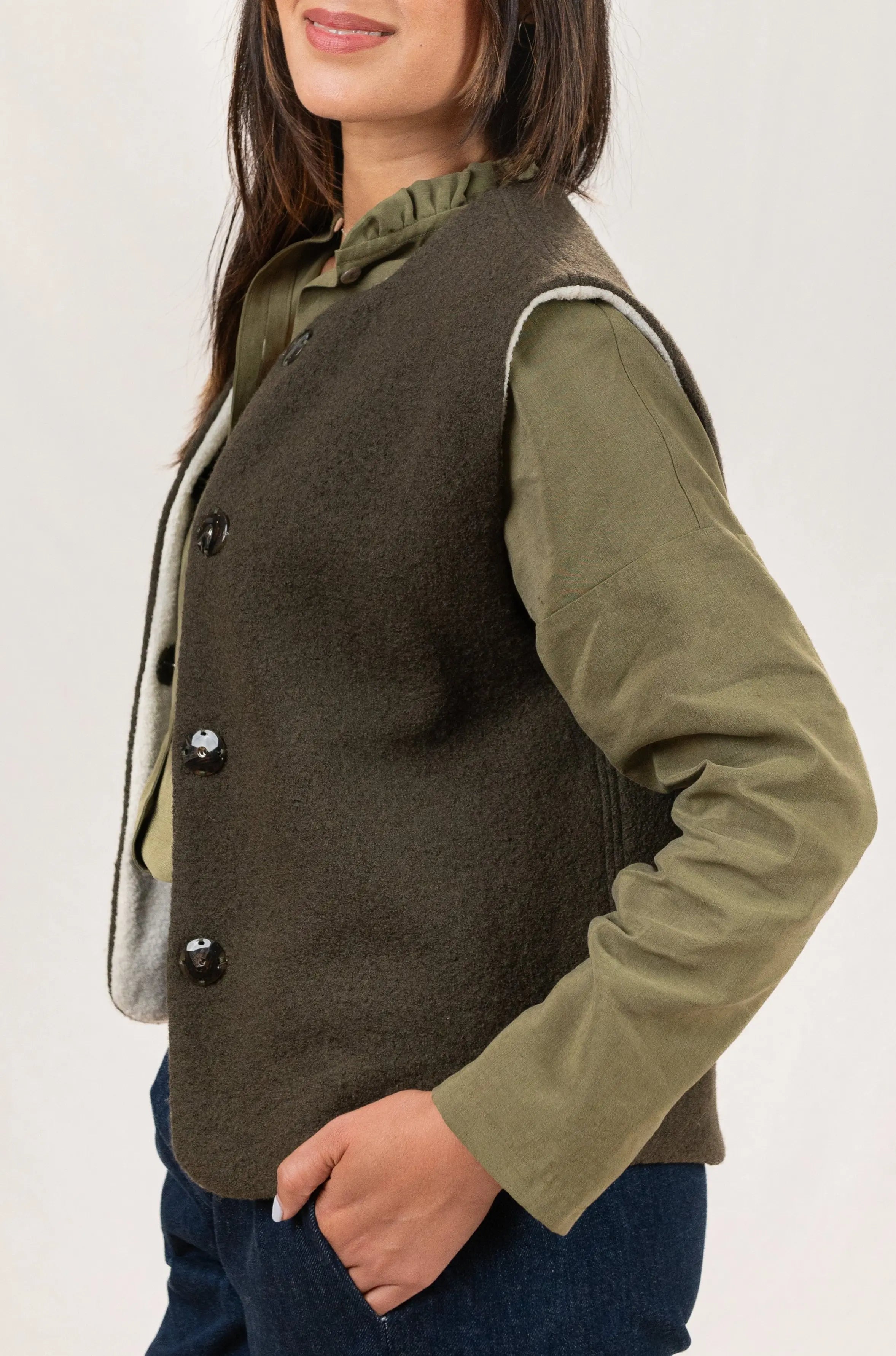 Gilet sans manches Ouessant réversible en laine bouillie vert forêt, vu de profil, porté avec une chemise Narcisse en lin et soie couleur kaki et un pantalon Sylène en denim bleu foncé.
