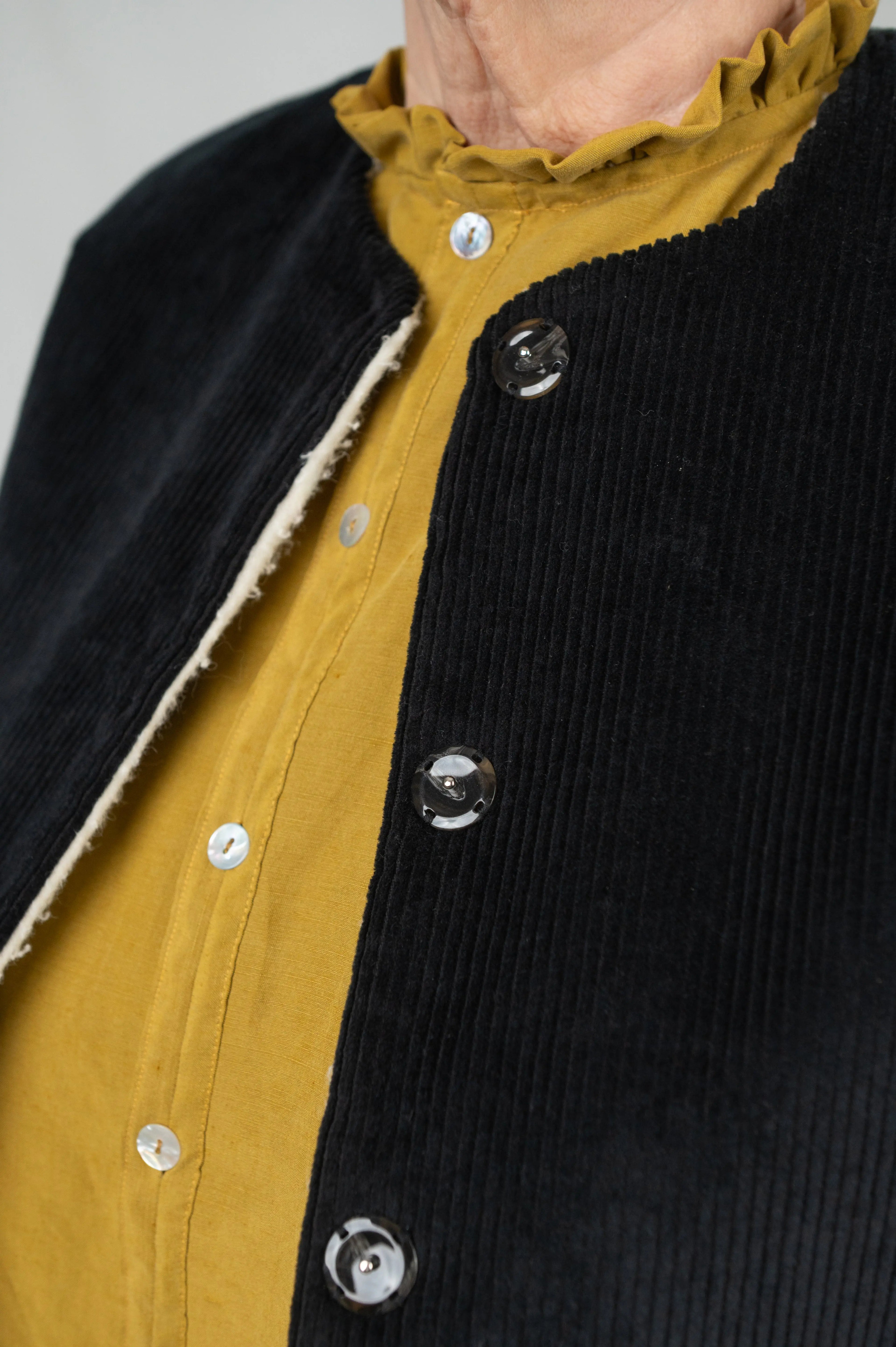 Gilet sans manches Ouessant en velours noir, détail des boutons effet corne, porté sur une chemise Narcisse en lin et soie moutarde.
