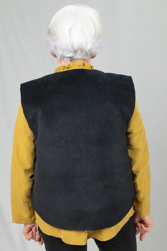 Gilet sans manches Ouessant réversible en velours noir, vu de dos, détail de la coupe dos recouvrant, porté sur une chemise Narcisse en lin et soie moutarde, chemise portée sortie du pantalon slim noir.