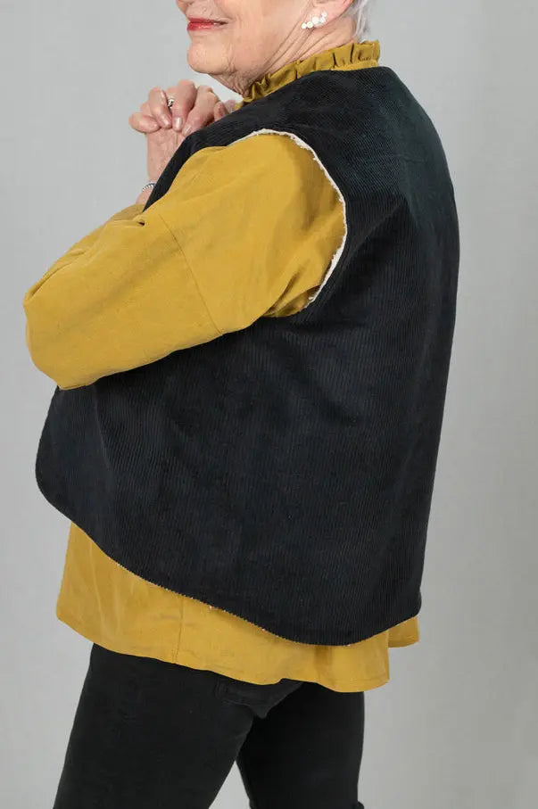 Gilet sans manches Ouessant en velours noir, vu de profil avec détail de la coupe dos recouvrant, porté sur une chemise Narcisse en lin et soie moutarde et un pantalon slim noir.