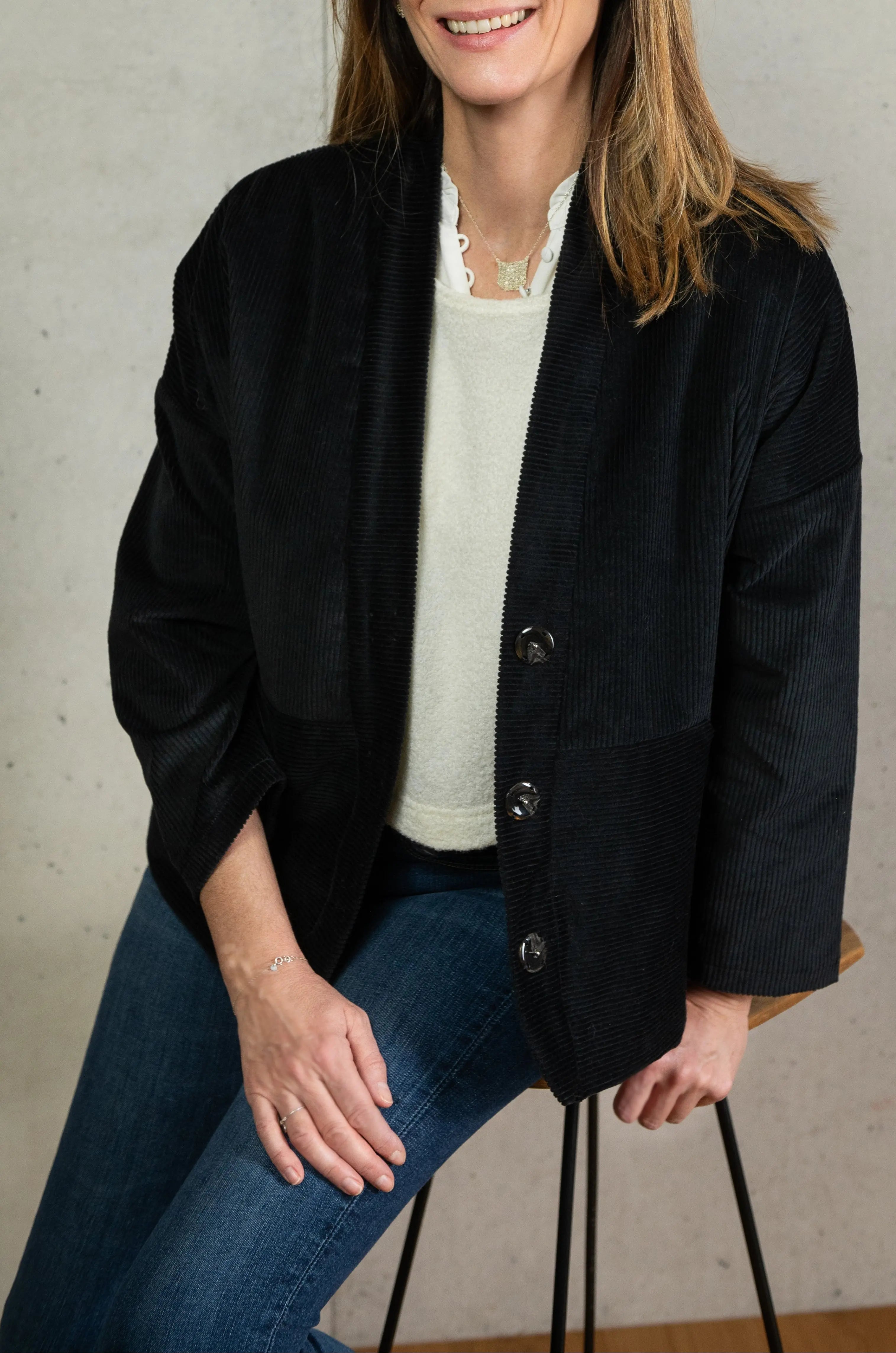 Manteau Cévennes coupe kimono en velours côtelé noir, vu de face porté avec un pull Mousse en jersey de laine crème et un bracelet et collier signé A.no.